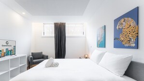 Une chambre d’hôtel moderne avec un grand lit, un canapé gris, un téléviseur posé sur un meuble blanc et deux œuvres d’art accrochées au mur.