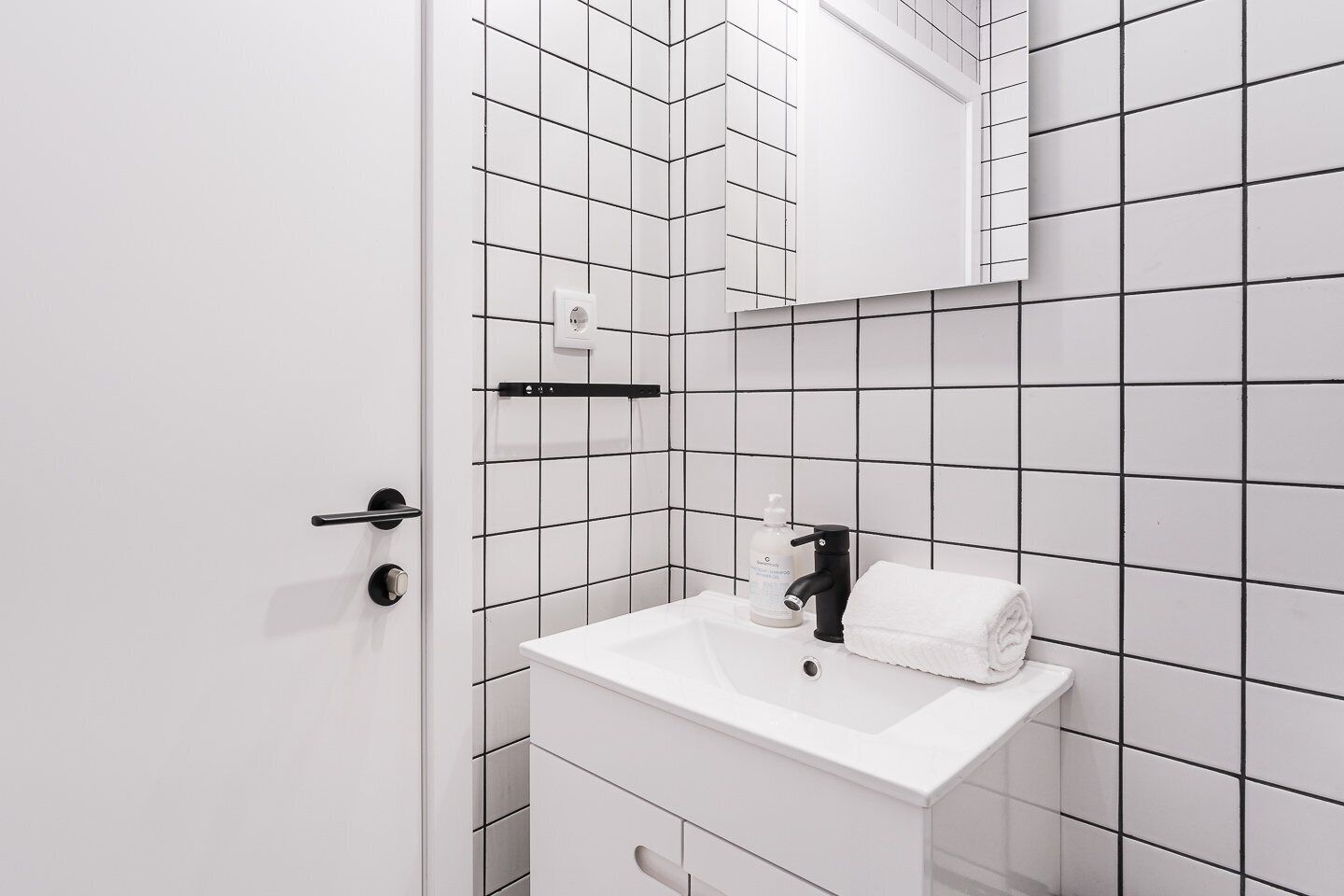A modern bathroom with a white sink, black faucet, and a mirror above it. There is a towel hanging on the wall and a soap dispenser on the sink.
