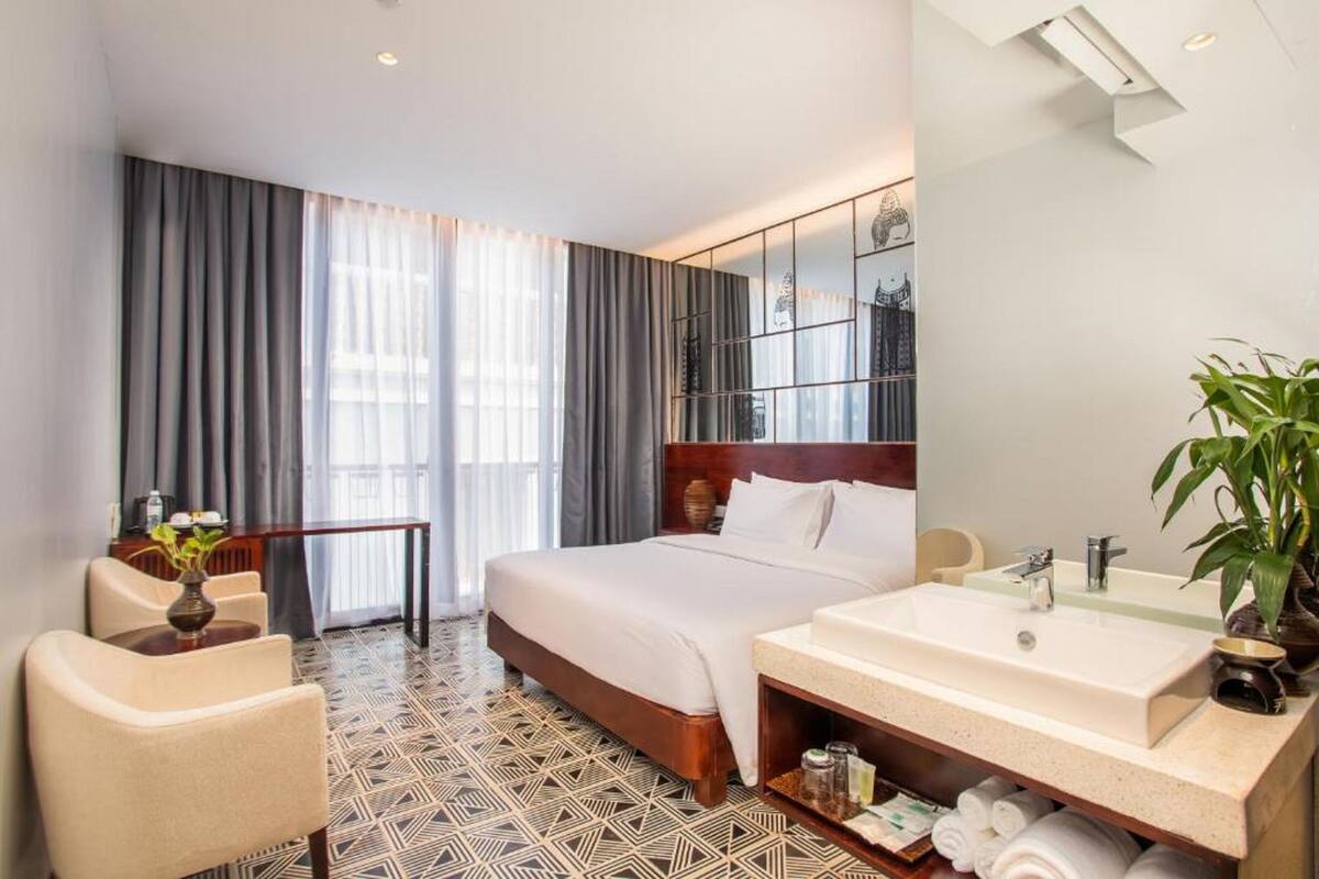 Habitación de hotel con cama, escritorio, silla, ventana grande con cortinas, baño con lavamanos y espejo, y una planta en maceta.