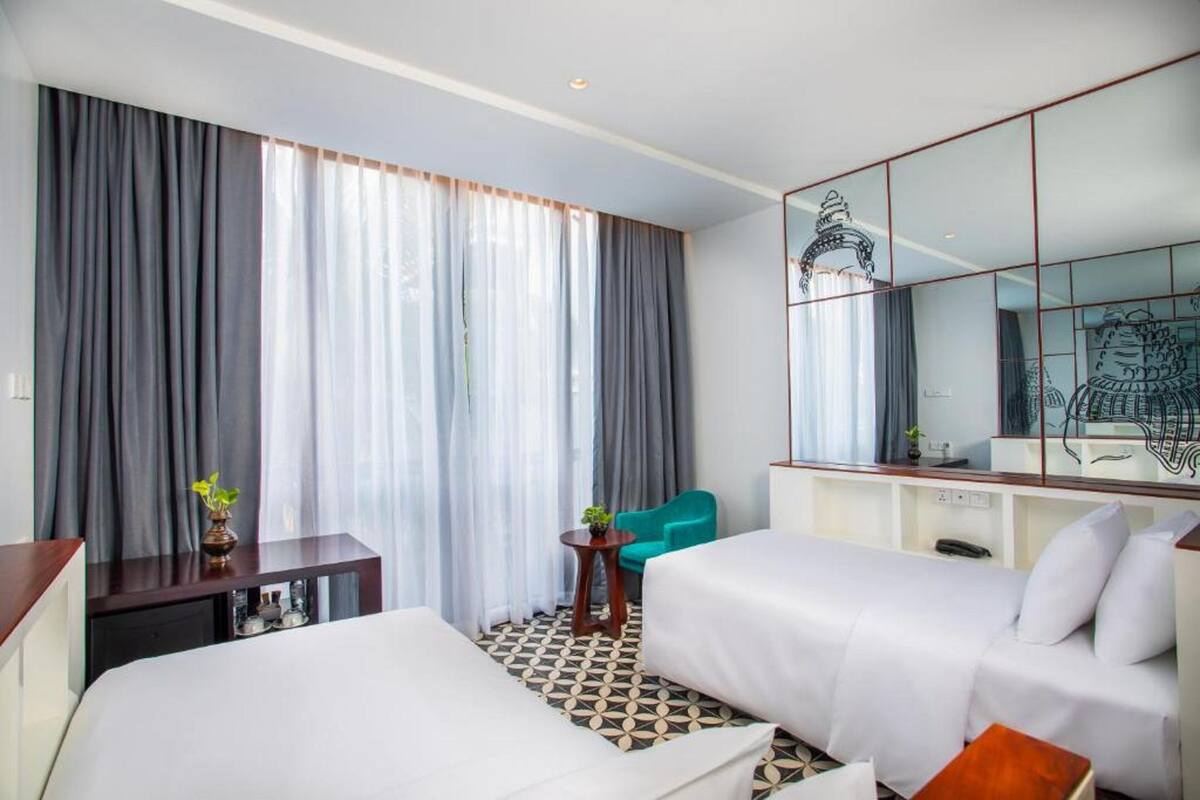Una habitación de hotel con dos camas, un escritorio, una silla, una mesita con una planta y un espejo grande.
