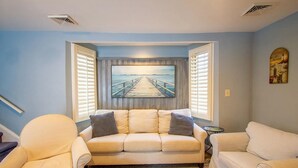 A living room with a beige sofa, two armchairs, a rug, a painting of a pier, and a small table.