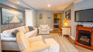 A living room with a white sofa, a white armchair, a small table with a lamp, a fireplace, and a TV.