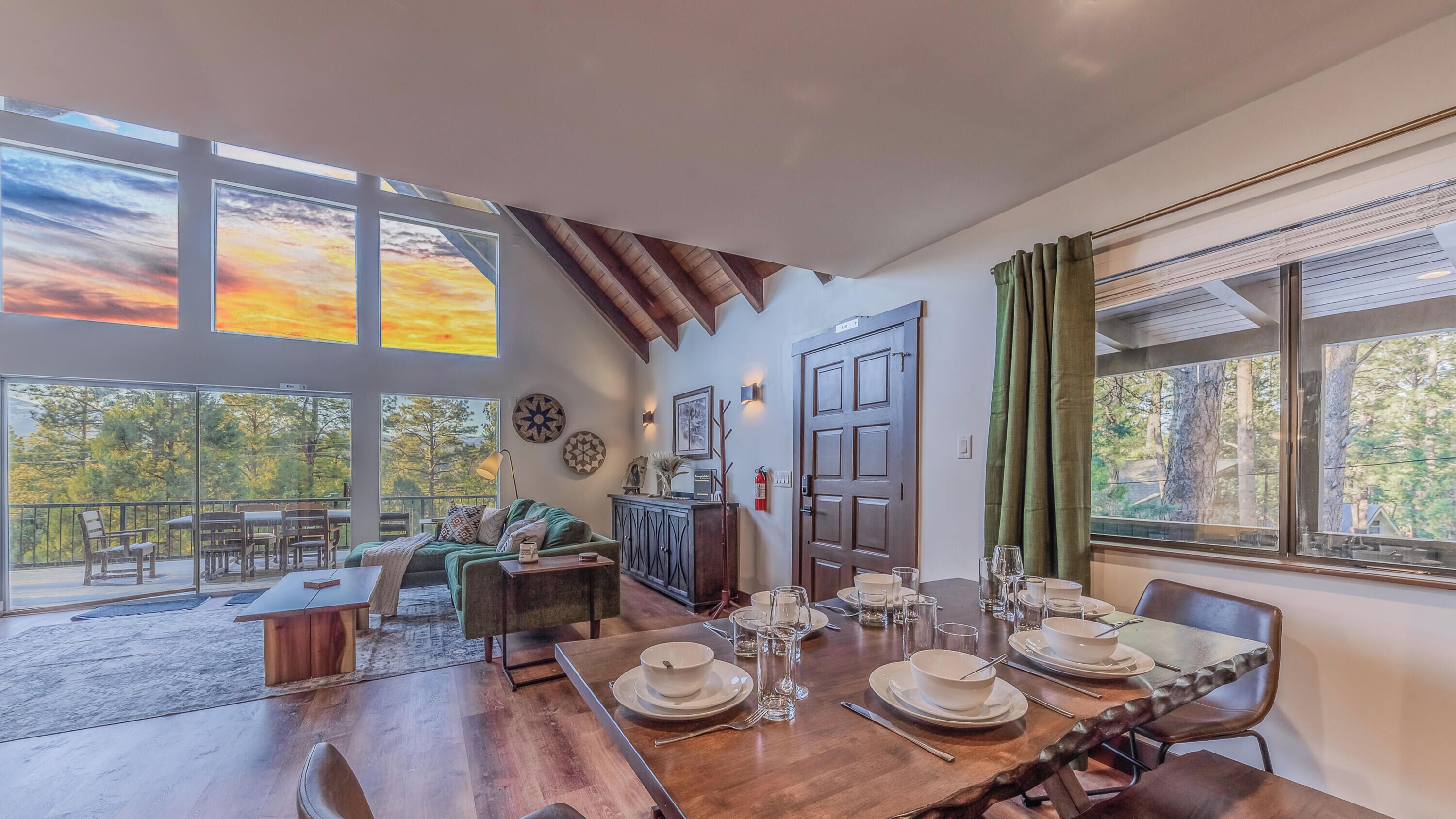 Une salle à manger spacieuse avec une table en bois dressée pour six personnes, de grandes fenêtres offrant une vue sur les arbres et un coin salon confortable avec une cheminée.