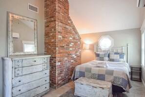 A bedroom with a brick wall, a white dresser, a bed with a quilt, and a window with a view.