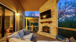 A wooden deck with wicker furniture, a stone fireplace, and a view of the surrounding trees. - Charming Creekside Escape: Pool, Putt-Putt, Game Room & Pet Friendly (Broken Bow)
