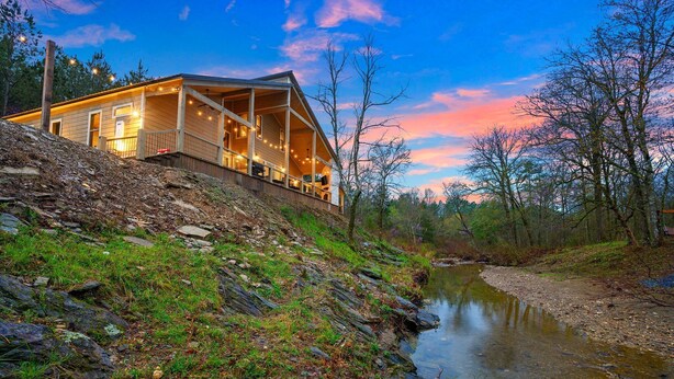 A cabin with a porch overlooking a river, surrounded by trees and a rocky landscape. - Charming Creekside Escape: Pool, Putt-Putt, Game Room & Pet Friendly (Broken Bow)