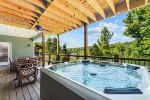 A wooden deck with a hot tub, outdoor seating, and a view of a lake surrounded by trees.