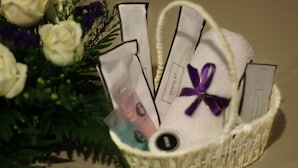 Un panier contenant des articles de soins dentaires et orné d’un ruban violet, entouré de roses blanches et de fleurs violettes.
