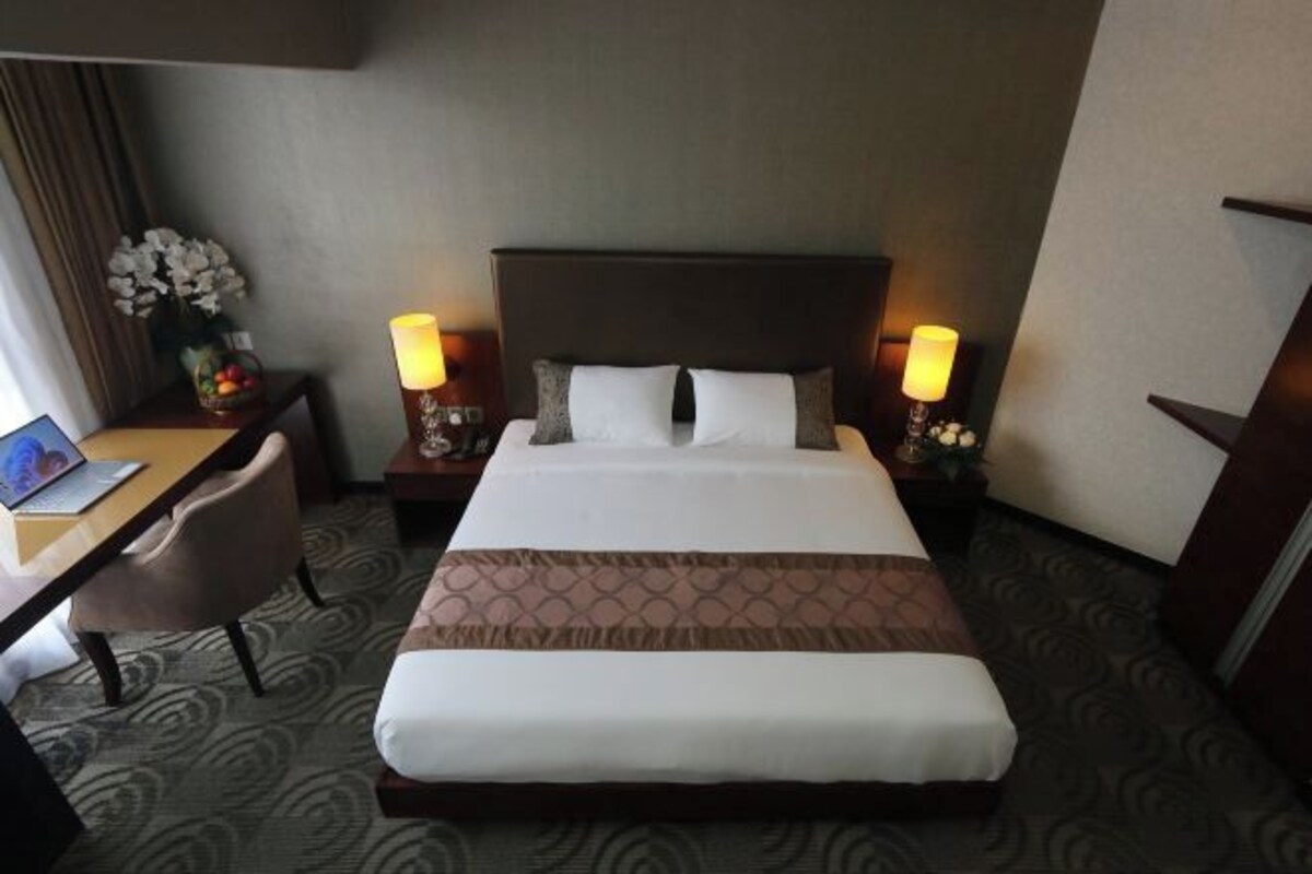 Een hotelkamer met een groot bed, twee nachtkastjes met lampen, een bureau met stoel, en een bloevas.