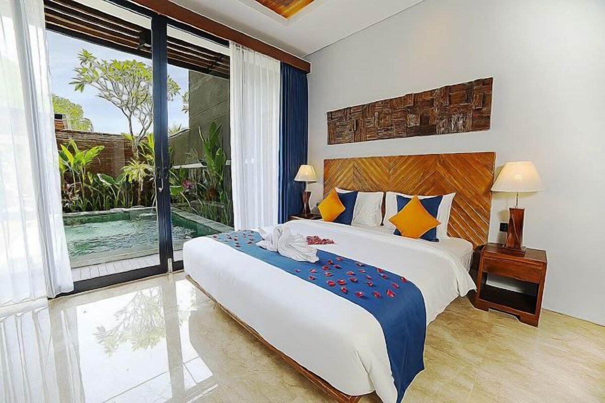 Habitación de hotel con una cama grande, cabecera de madera, dos lámparas de noche, vista a una piscina y puerta corrediza al exterior.