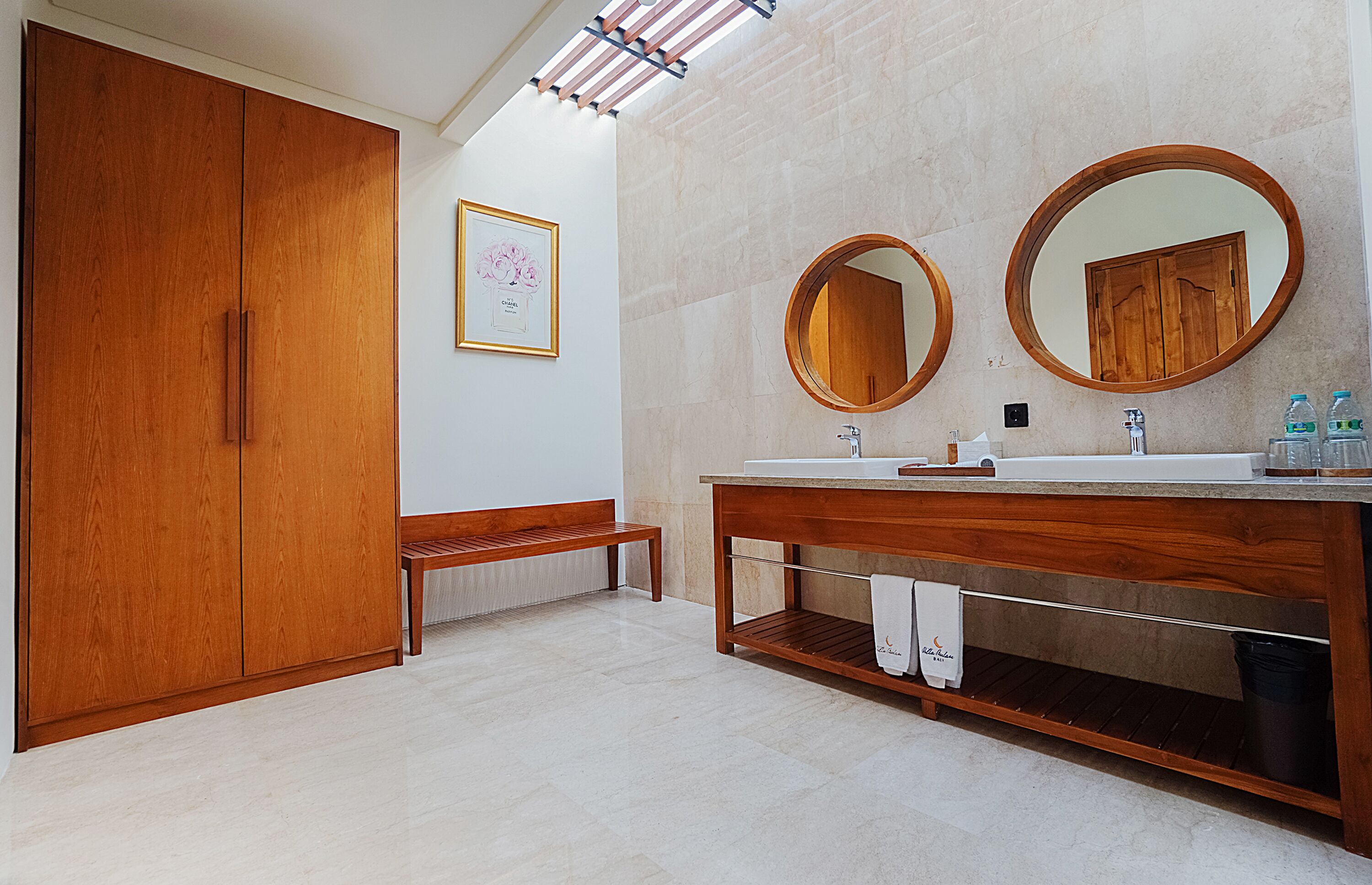 A modern bathroom with a wooden vanity, two round mirrors, a bench, and a framed artwork on the wall.