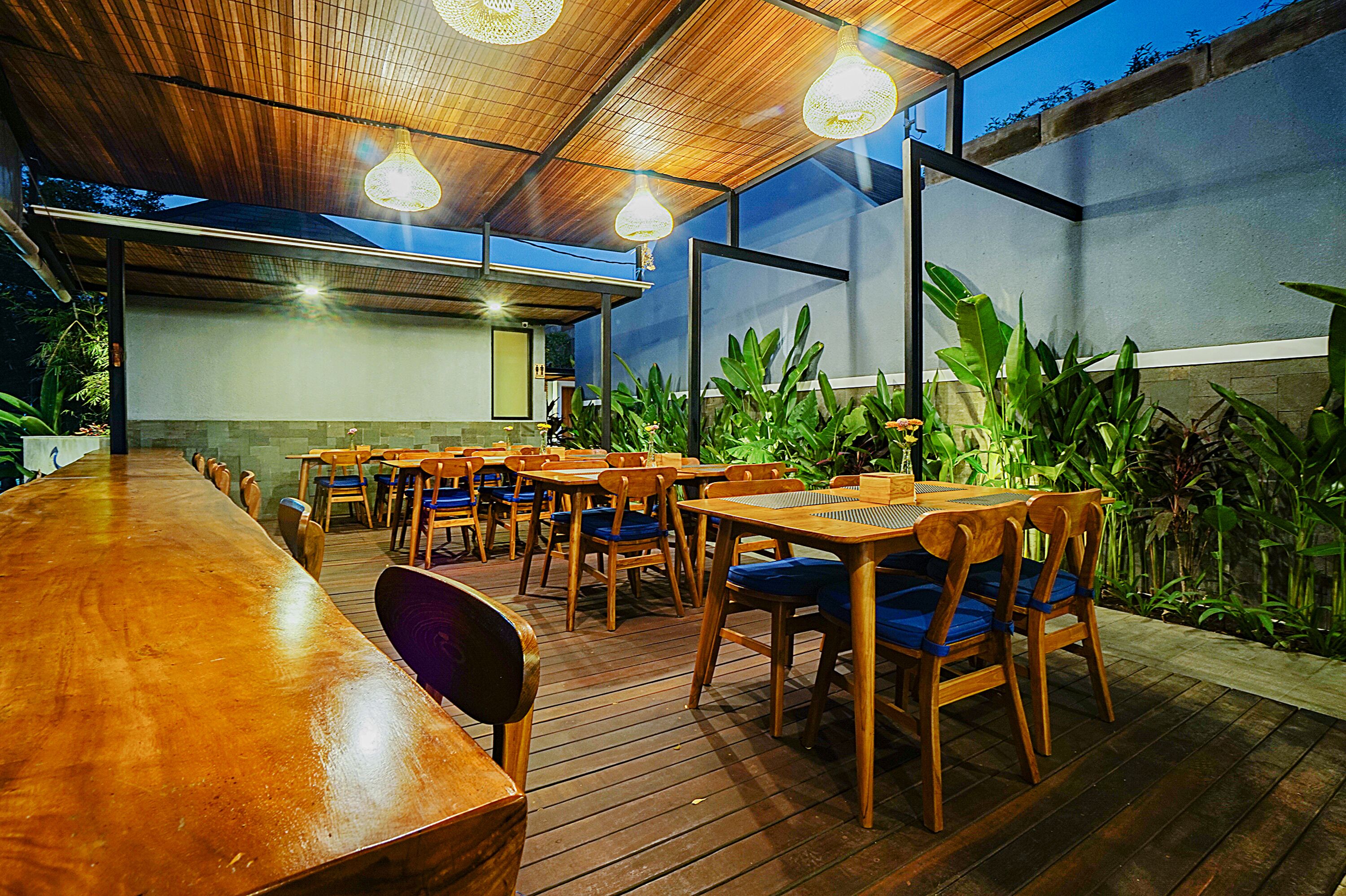 An outdoor dining area with wooden tables and chairs, surrounded by plants.