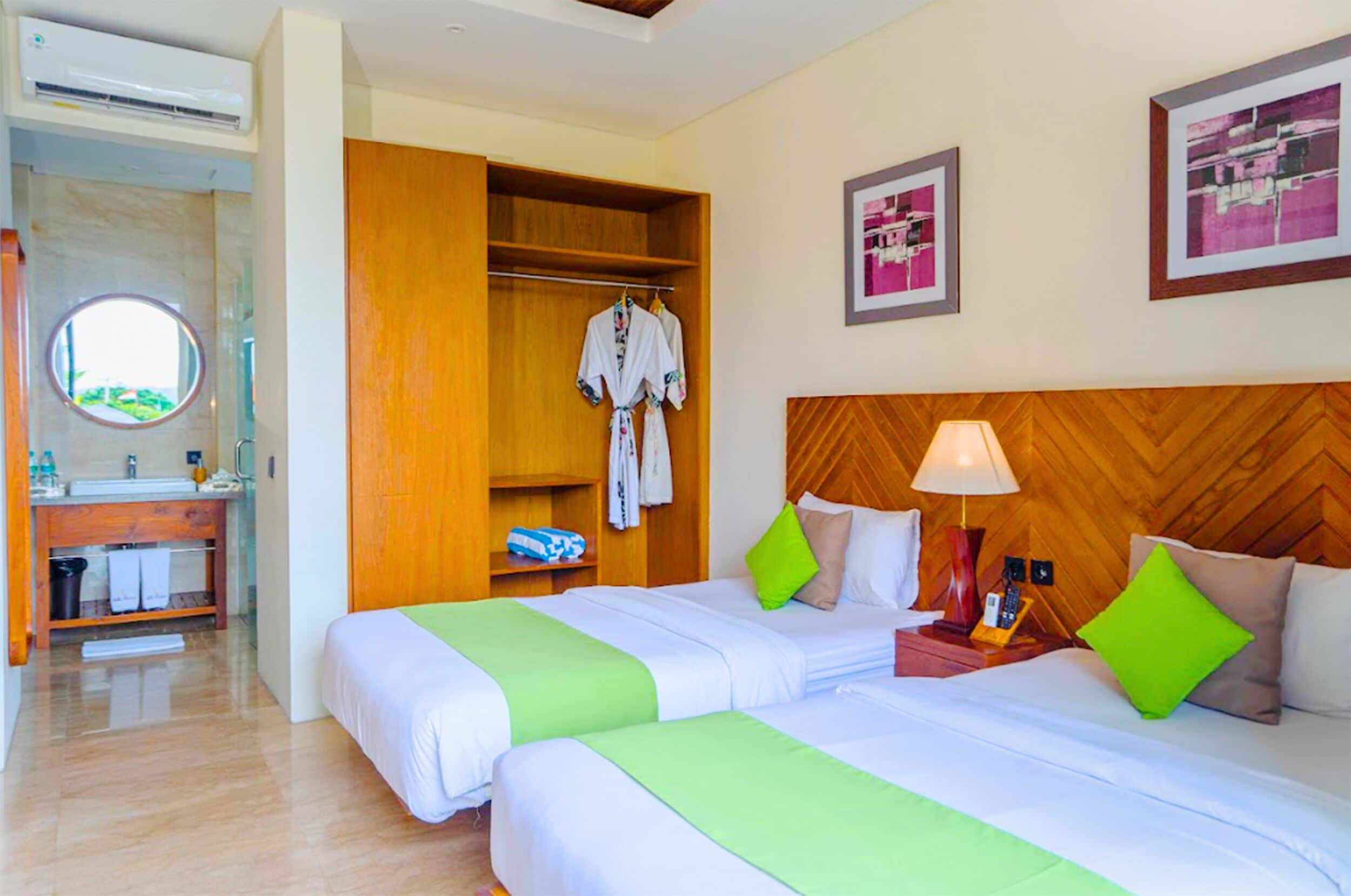 A hotel room with two beds, a wooden headboard, a bedside table with a lamp, and a bathroom with a round mirror and sink.