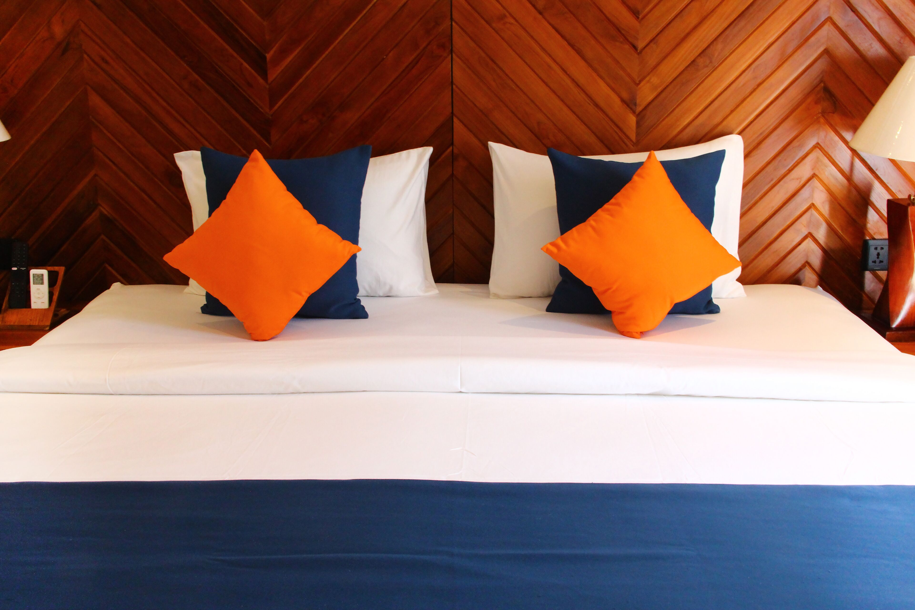 Una cama con ropa de cama blanca y azul, dos almohadas naranjas y un cabecero de madera con diseño de espiga.