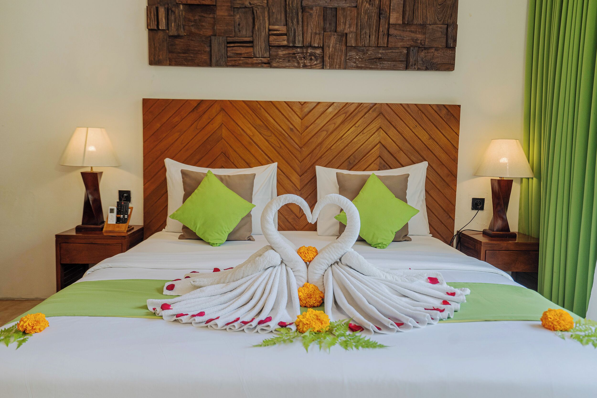 Habitación de hotel con una cama decorada con toallas formando un corazón, almohadas verdes y un cabecero de madera.