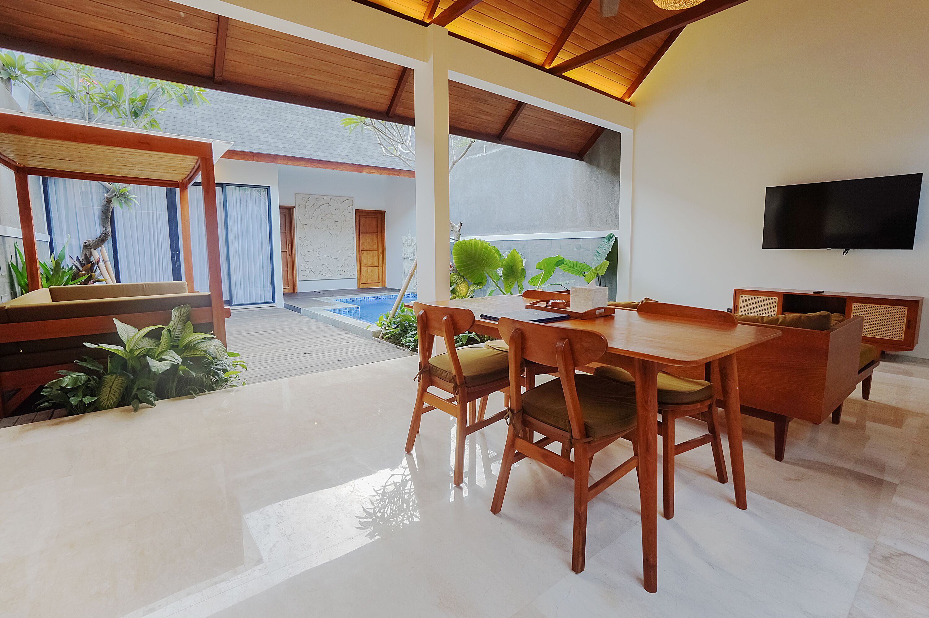 A modern dining area with a wooden table and chairs, a flat-screen TV mounted on the wall, and a view of a swimming pool and lush greenery.