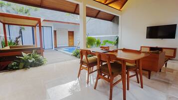 A modern dining area with a wooden table and chairs, a flat-screen TV mounted on the wall, and a view of a swimming pool and lush greenery.
