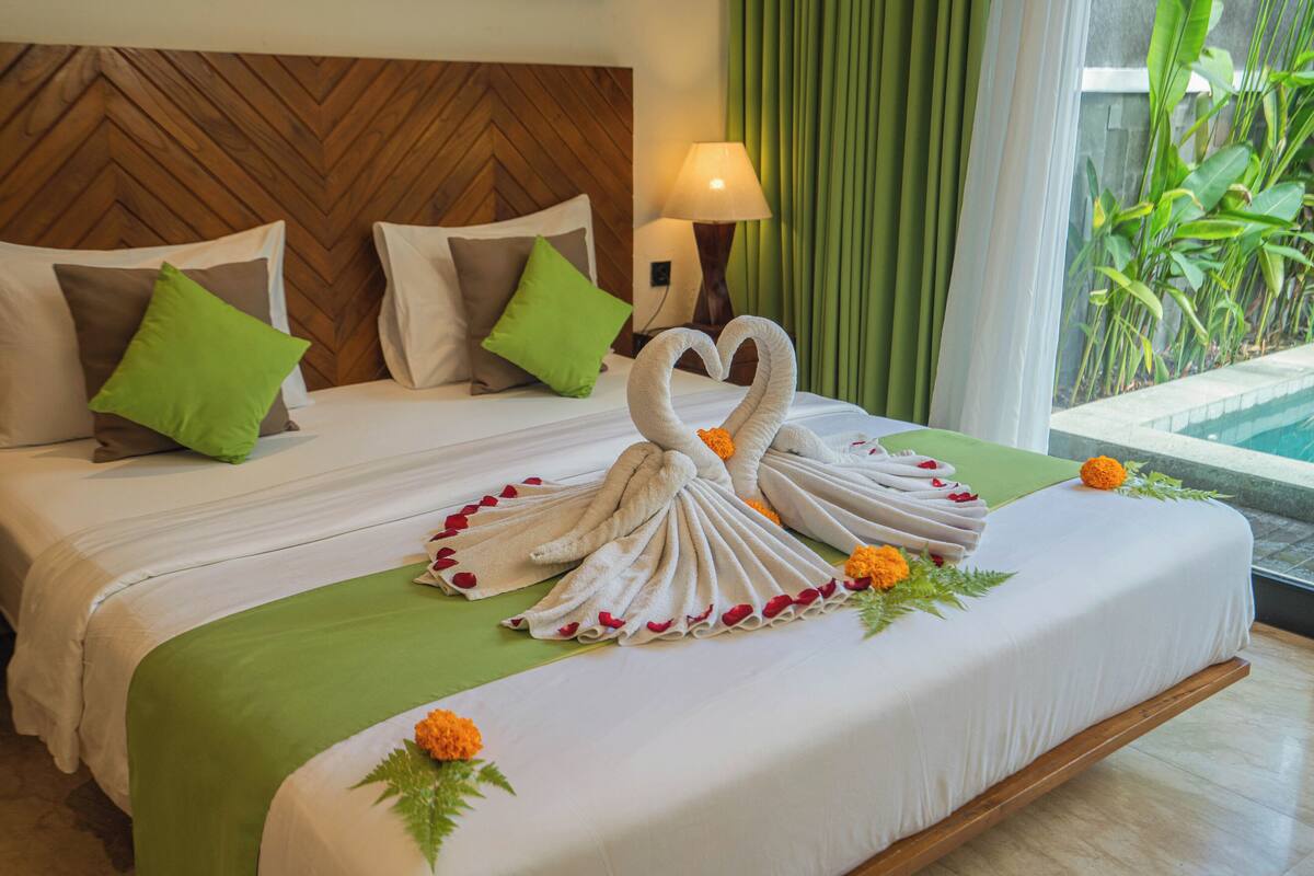 A neatly made bed with a green bedspread, white pillows, and a heart-shaped towel arrangement on the bed.