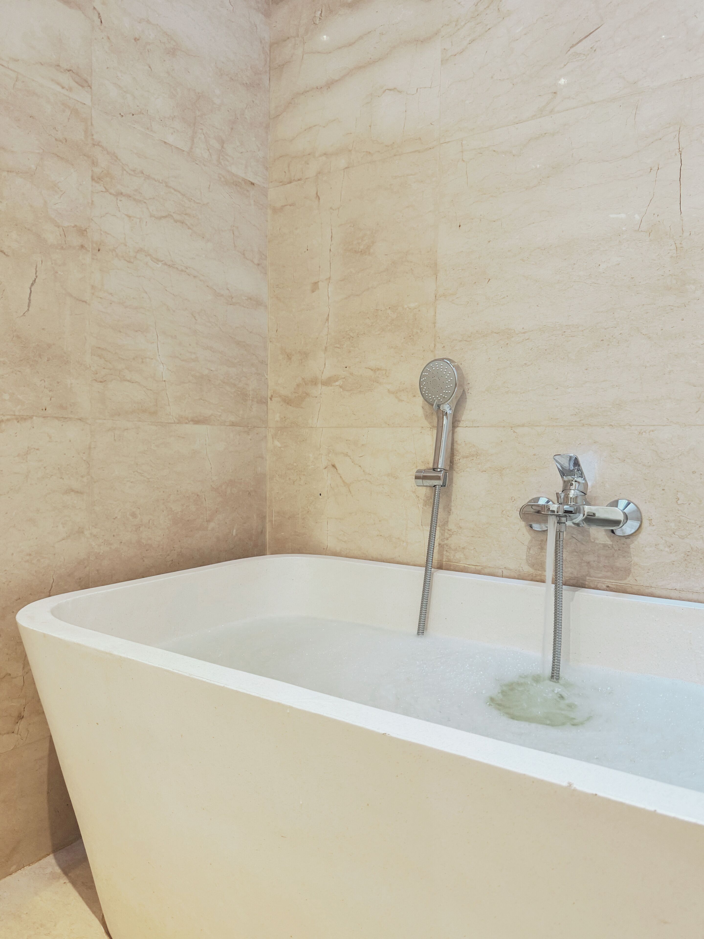 Una bañera blanca independiente con cabezal de ducha manual y cabezal de ducha fijo, frente a una pared de mármol.