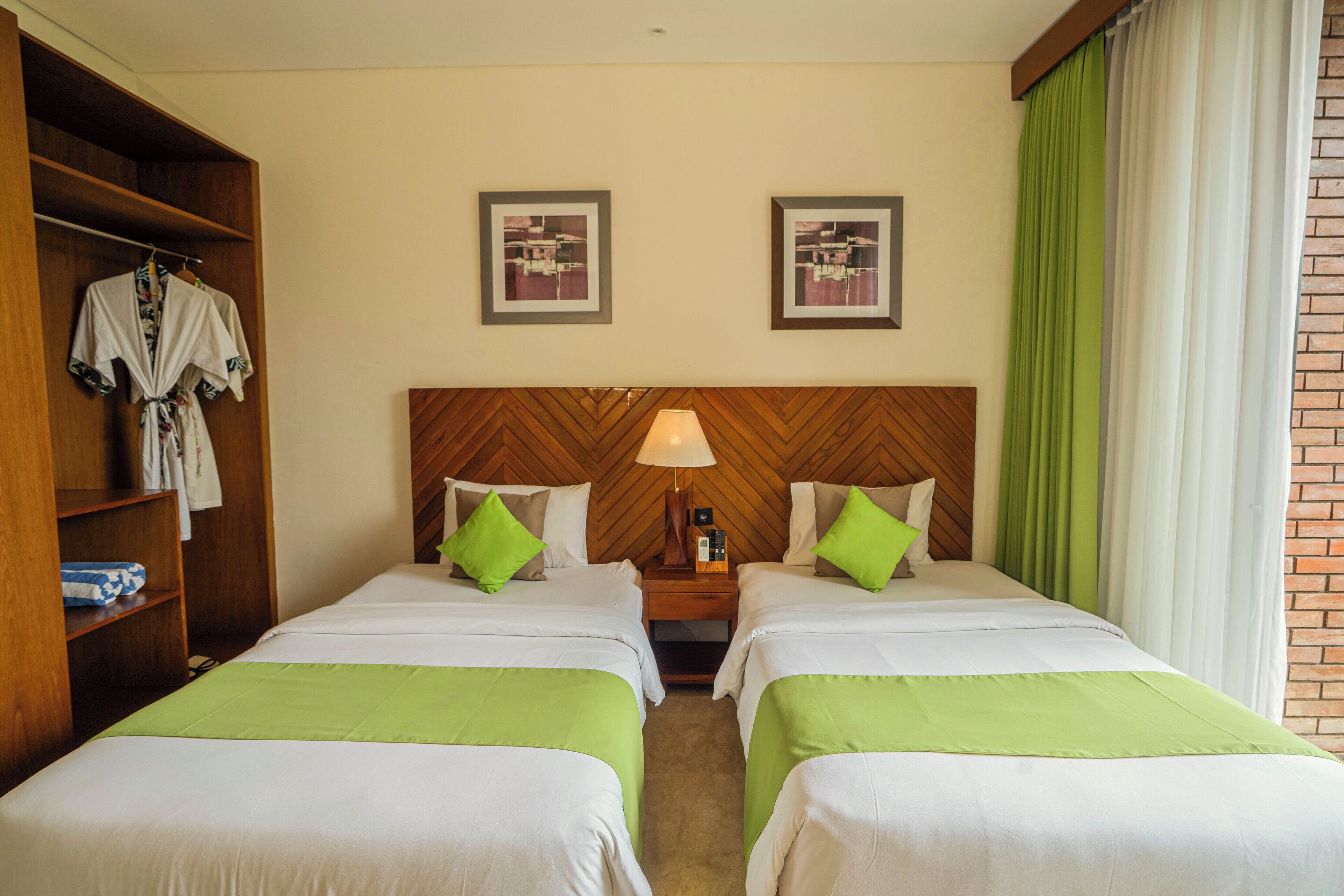 A hotel room with two beds, a wardrobe, a nightstand with a lamp, and two framed pictures on the wall.