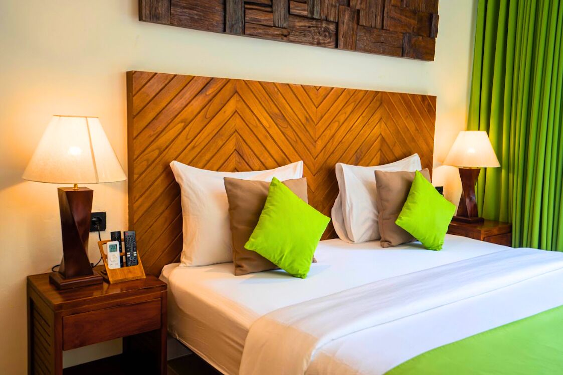 A hotel room with a wooden headboard, two bedside lamps, a bedside table with a phone and remote controls, and two green pillows on the bed.