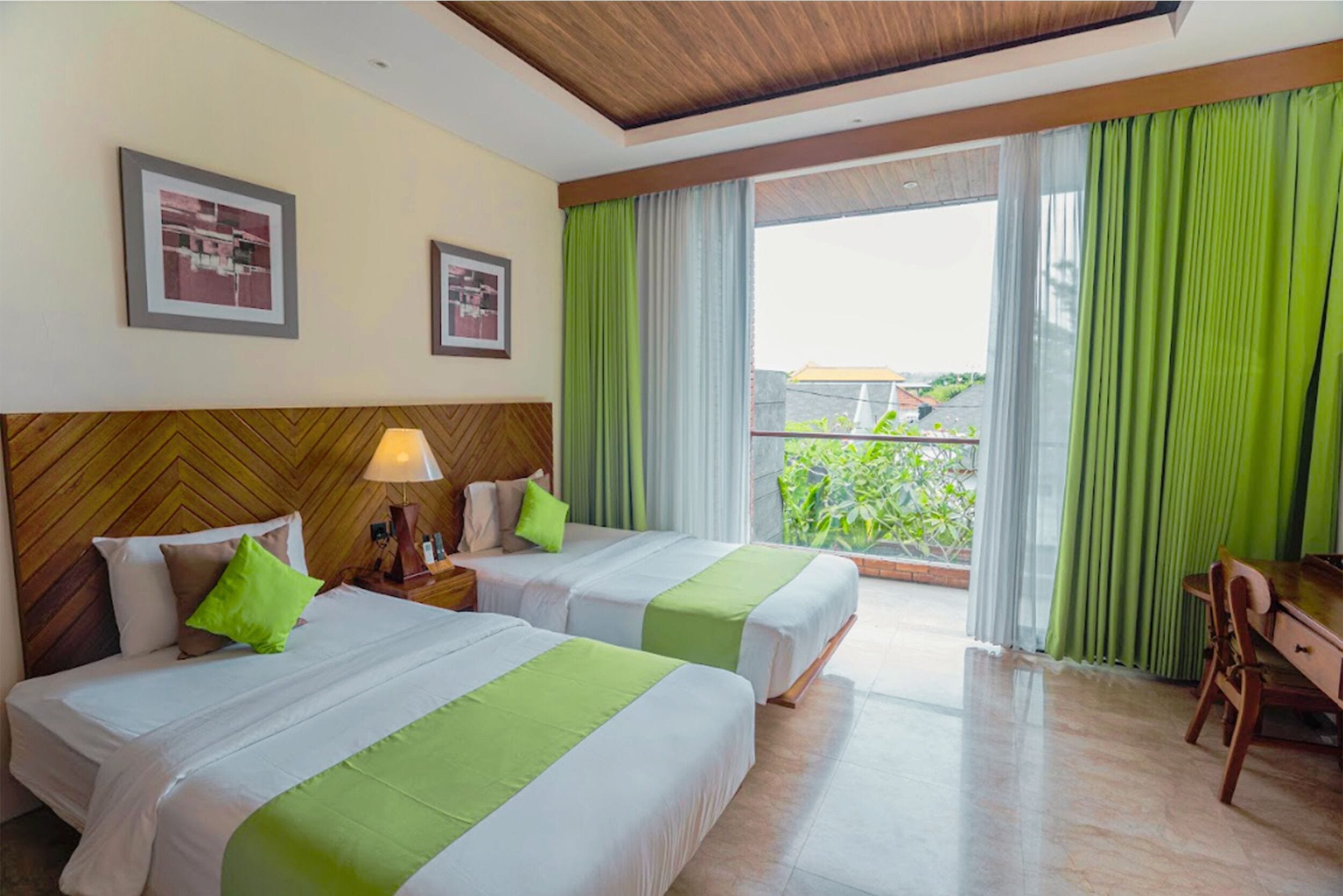 Quarto de hotel com duas camas, uma cabeceira de madeira, uma escrivaninha e uma varanda com cortinas verdes.