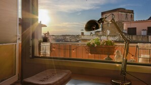 Una habitación con escritorio, computadora portátil, ventana con vista a edificios, lámpara y cojín con texto.