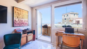 Una habitación con escritorio, silla, televisión, estantería y un amplio ventanal con vistas a edificios.