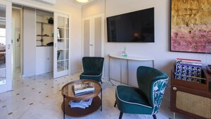 Una sala de estar con televisor, dos sillones verdes, una mesa de centro con revistas, una mesita auxiliar y una estantería.