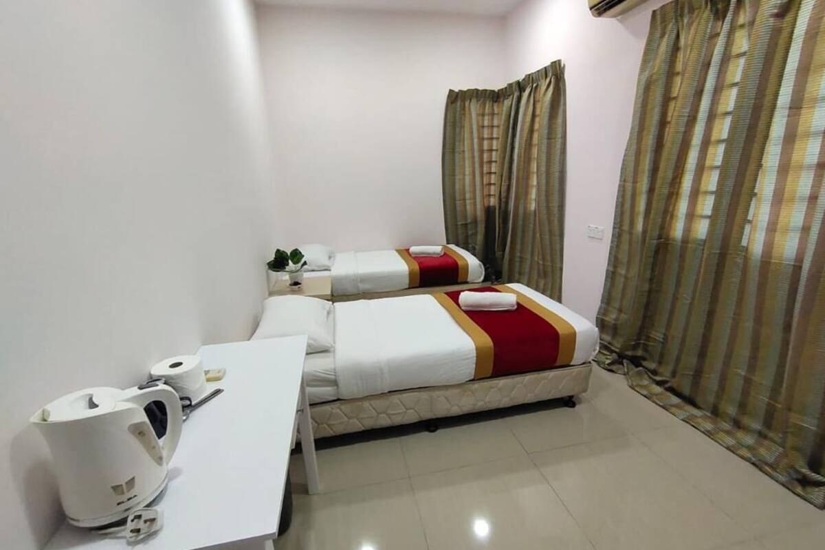 A hotel room with two beds, a white table with a kettle and a roll of toilet paper, and a window with curtains.