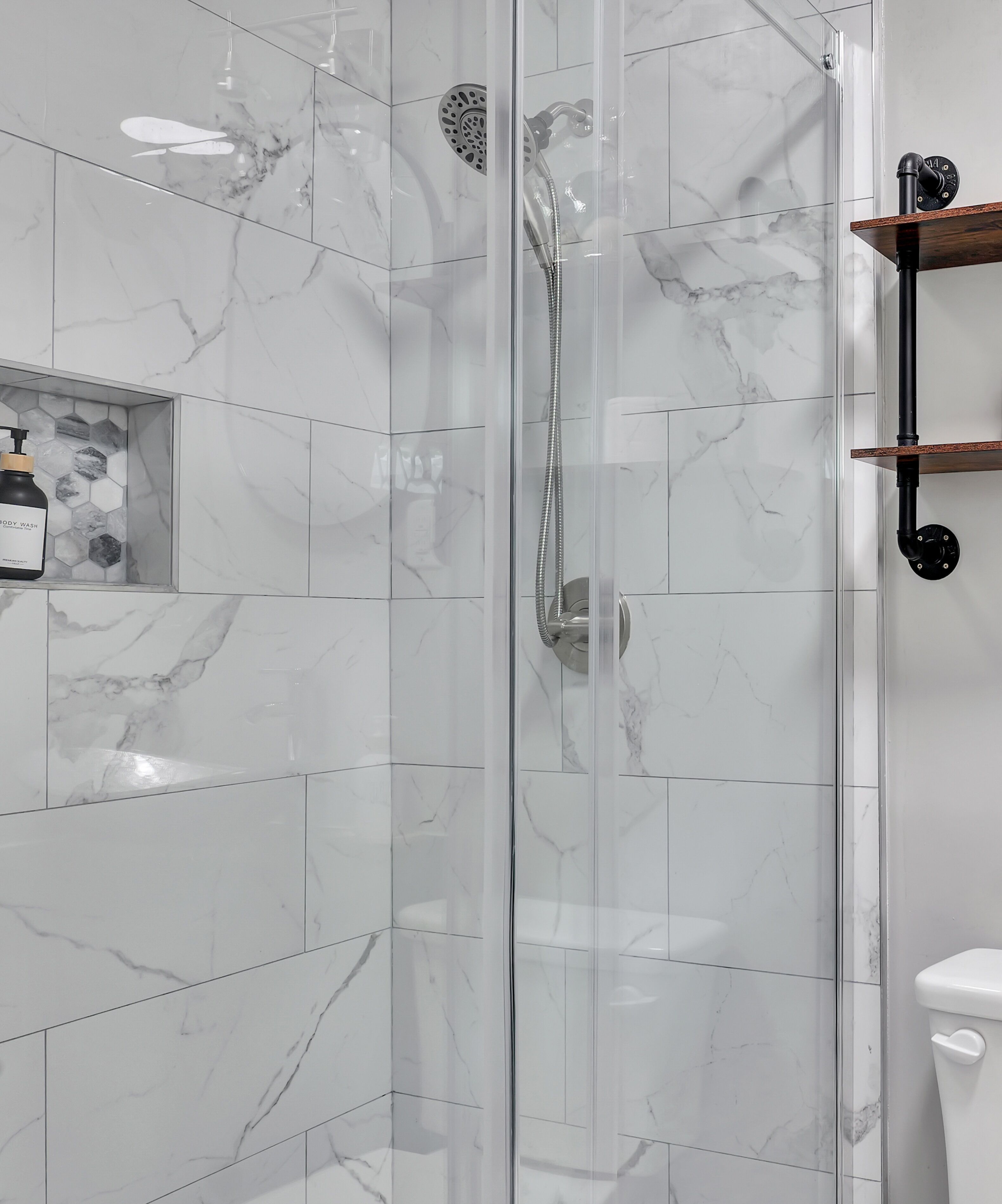 Une salle de bain moderne avec une douche à parois de verre, des carreaux de marbre et une étagère en bois où sont rangés les articles de toilette.