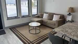 Ein modernes Wohnzimmer mit einem Ecksofa, einem runden Couchtisch und einem Esstisch mit Stühlen. Große Fenster bieten Blick auf eine verschneite Landschaft.