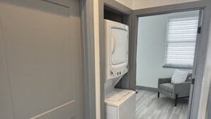 Ein Waschraum mit Waschmaschine und Trockner, ein grauer Stuhl und ein Fenster mit Jalousien.