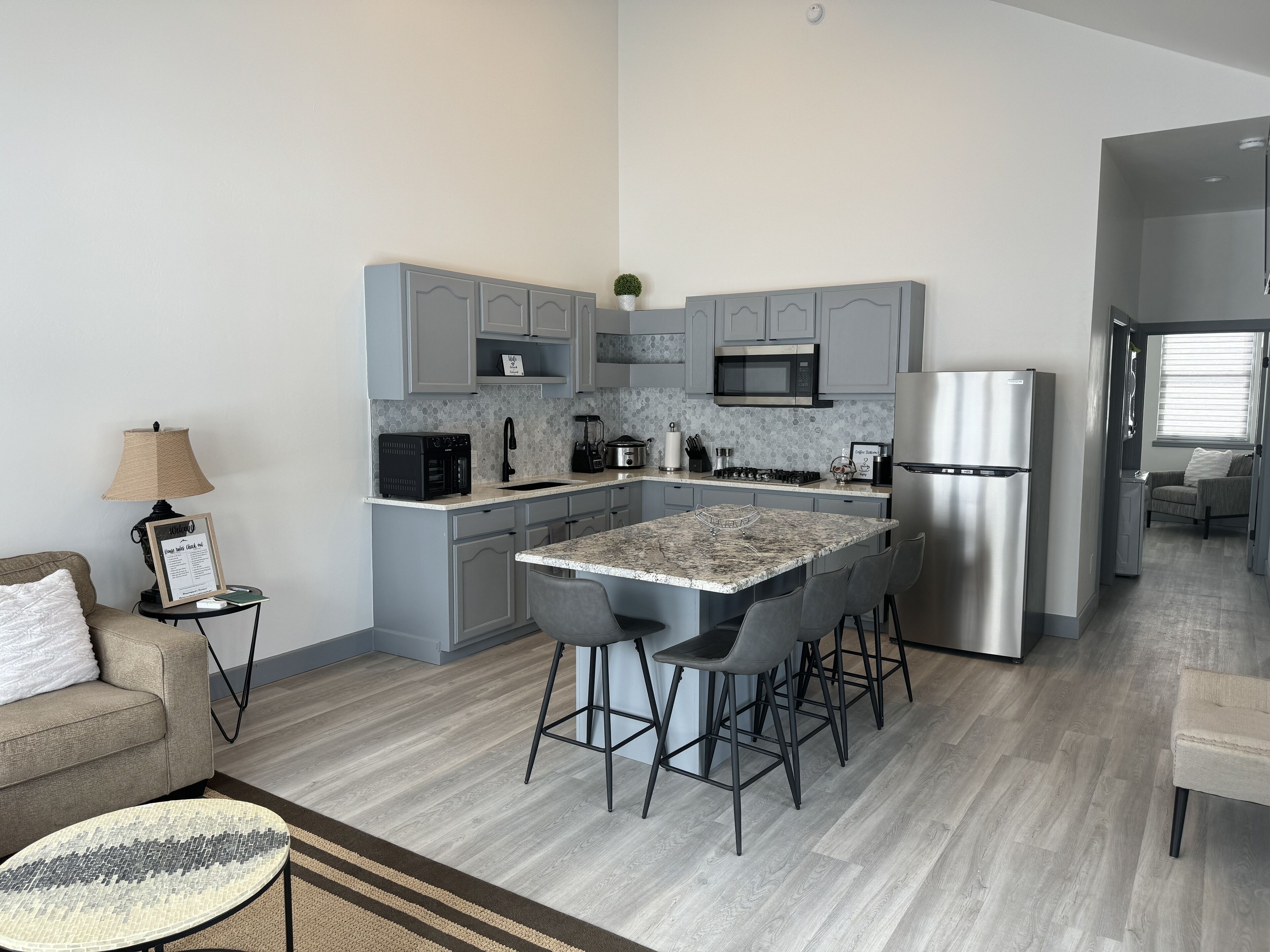 A modern kitchen with grey cabinets, a central island with bar stools, a built-in microwave, and a stainless steel refrigerator.