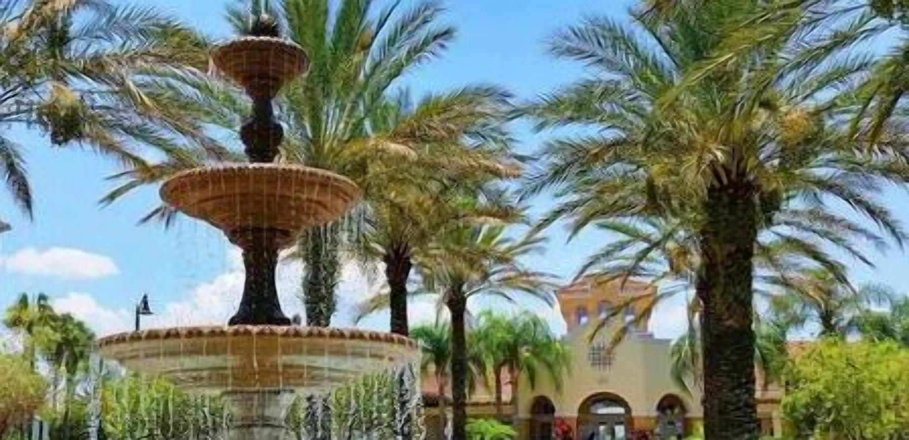 A multi-tiered fountain surrounded by palm trees and a building with arches in the background.