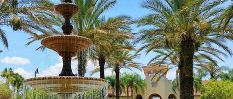 A multi-tiered fountain surrounded by palm trees and a building with arches in the background.