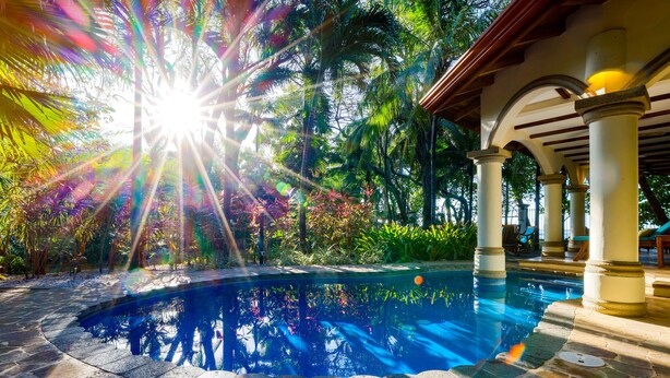 A pool area with stone flooring, palm trees, and a covered seating area with columns.