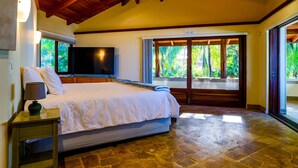 A bedroom with a large bed, a TV, and a window with a view of greenery.