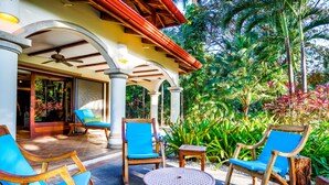 An outdoor seating area with wooden chairs and a round table, surrounded by lush tropical plants and a covered patio.