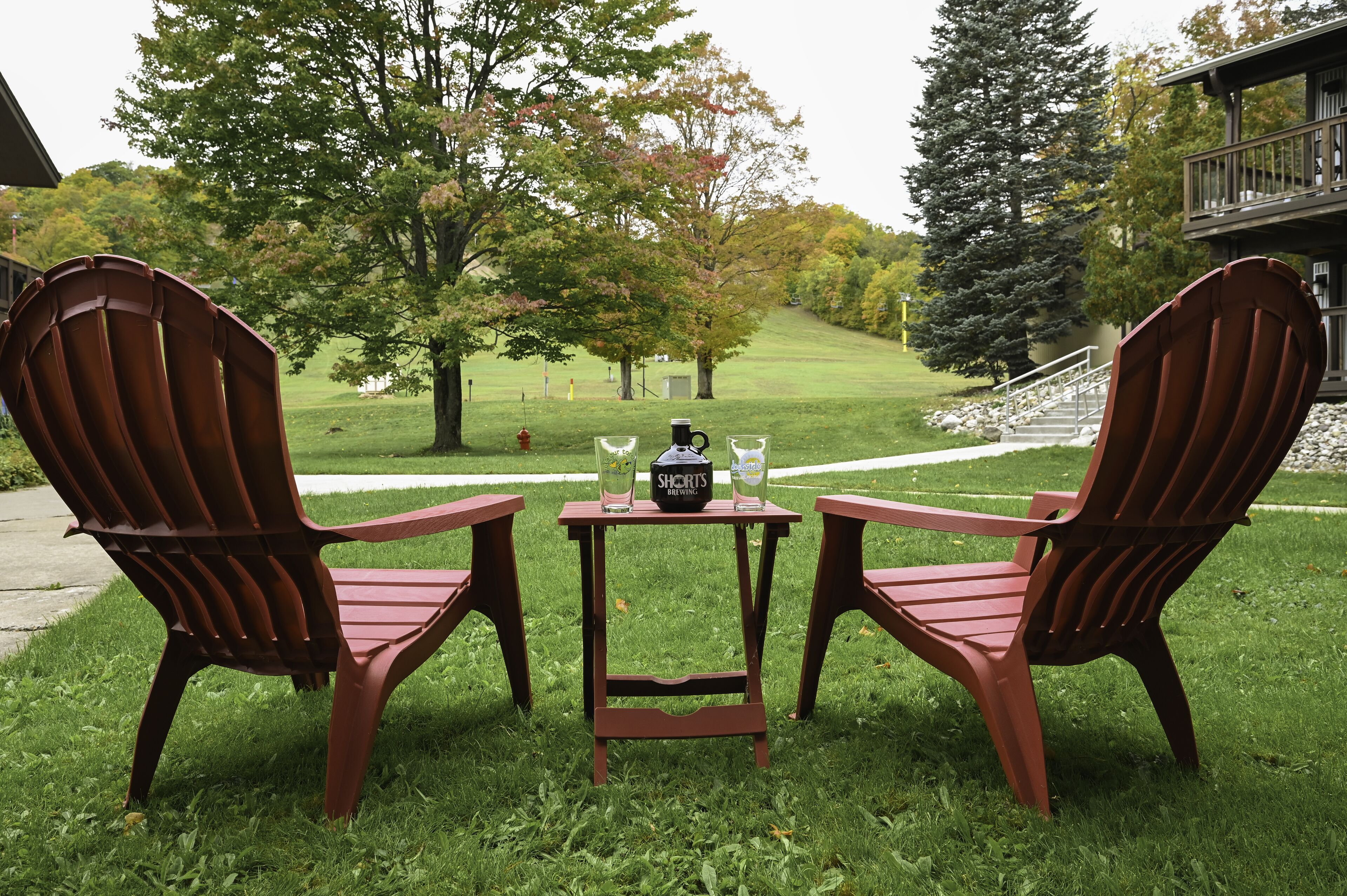 Deux fauteuils Adirondack en bois, avec une petite table placée entre eux, sur laquelle se trouvent deux bouteilles et une tasse à café.