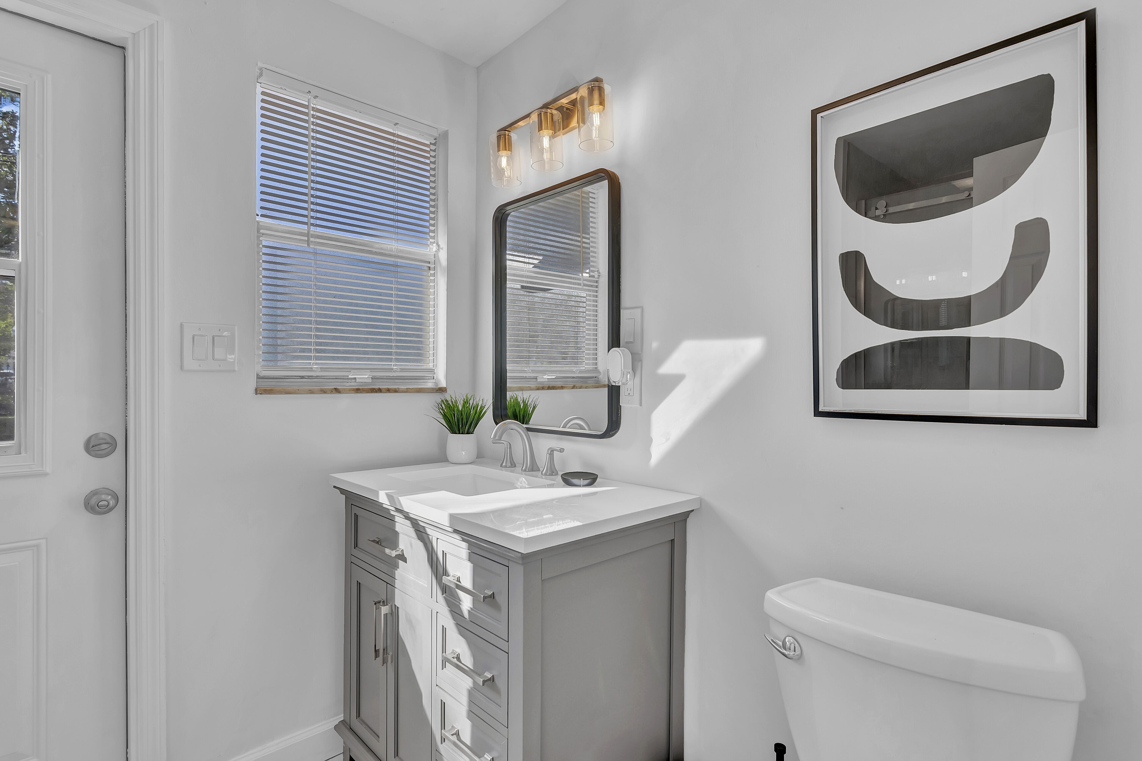 A bathroom with a white sink, a mirror, a toilet, and a window with blinds.