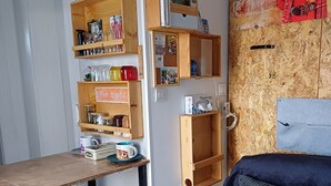 A compact room with a wooden table, two stools, a bed with a dark headboard, and a wall-mounted shelf with various items. - 10-square-meter studio apartment + 18-square-meter terrace + Pool + Private parking  (Quissac)