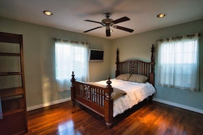 Quarto com uma cama de madeira com dossel, um ventilador de teto e duas janelas com cortinas.
