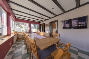 Un comedor con una mesa y sillas de madera, suelo de baldosas y un gran ventanal con persianas.