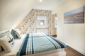 A bedroom with a bed, striped pillows, a floral-patterned wall, a window, and a painting. - Apartment "Kleiner Priel" with Wi-Fi (Wurster Nordseeküste)