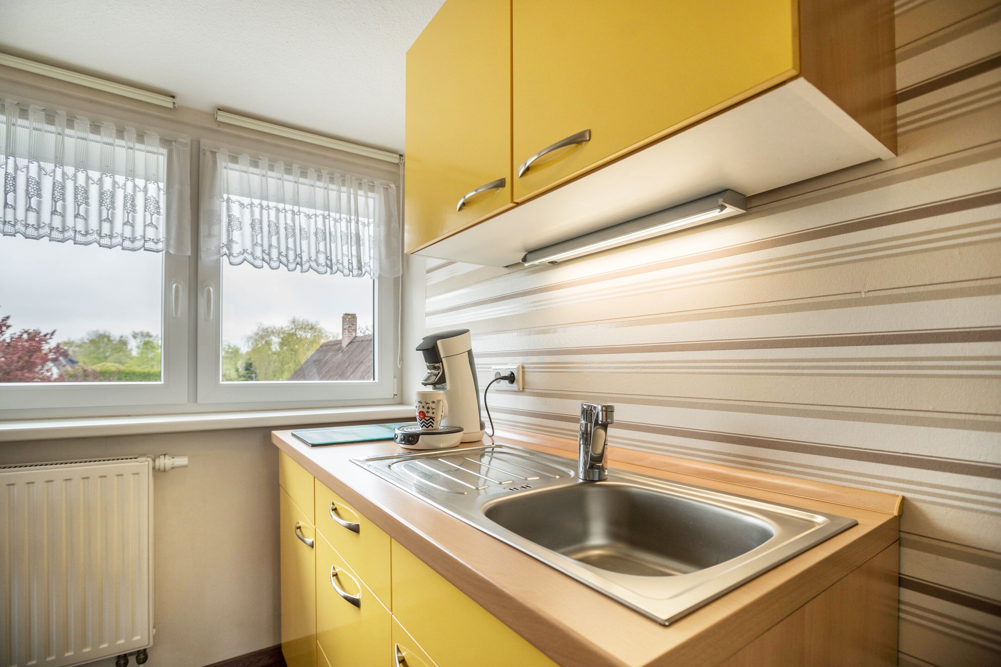A kitchen with yellow cabinets, a stainless steel sink, a window with curtains, and a radiator.