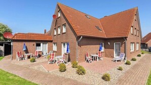 Ein zweistöckiges Backsteingebäude mit geneigtem Dach, zahlreichen Fenstern und einer gepflasterten Terrasse mit Außenbestuhlung.