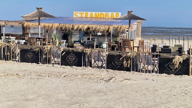 Ein an der Küste gelegenes Lokal namens 'STRANDRAP' mit einem Reetdach, Außenplätzen und Blick auf den Ozean.