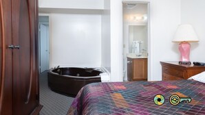 Quarto de hotel com uma cama grande, um guarda-roupa de madeira, um banheiro com banheira e um abajur rosa sobre uma mesinha de cabeceira.