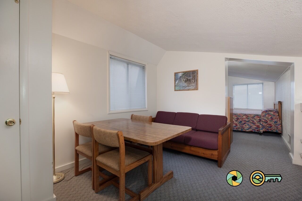 Un espacio habitable pequeño con una mesa de comedor y sillas, un sofá, una ventana con persianas y un cuadro pequeño en la pared.